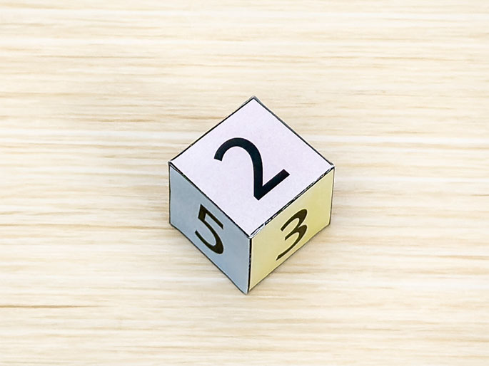 TwA completed, hand-made paper cube sits on a light wood-grain surface. The visible sides display the numbers 2, 3, and 5, each on a different pastel-colored square. This image shows the finished result of assembling a paper cube net.
