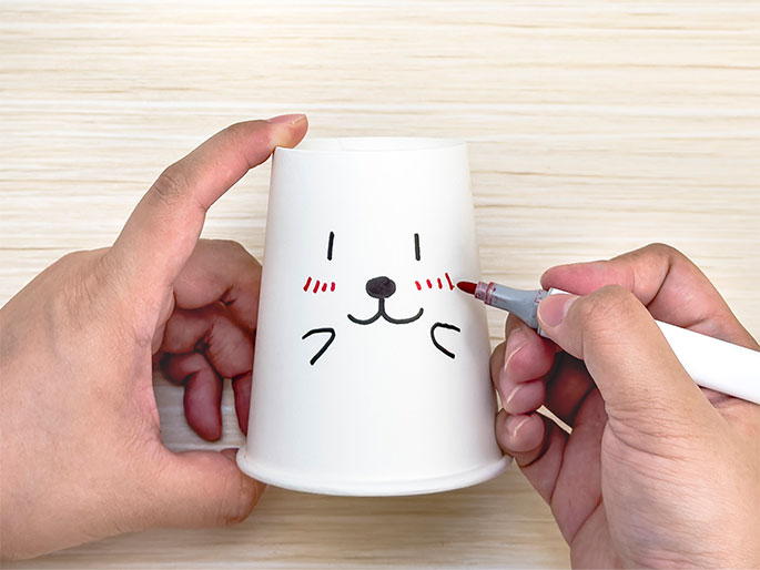 Two hands are decorating a white paper cup on a light wooden table. One hand holds the cup while the other draws rosy cheeks on a simple, cartoon animal face using a red marker. The face features short vertical lines for eyes, a small, rounded triangle for a nose, and curved lines for a smiling mouth. The image depicts a crafting activity, personalizing a paper cup with a cute design.