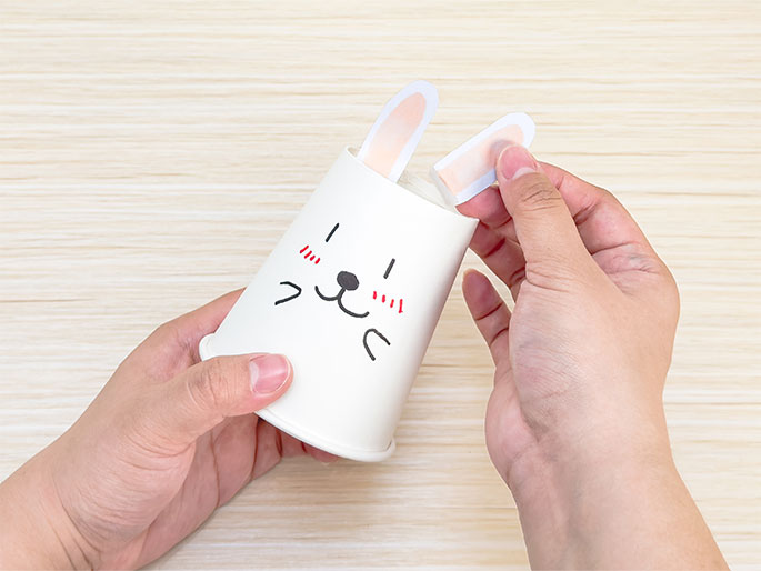 Two hands are assembling a paper cup craft on a light wooden table. One hand holds the white cup, which has a cute animal face drawn on it with rosy cheeks, simple eyes, a nose, and a smiling mouth. The other hand is attaching a pre-colored pink and white paper rabbit ear to the inside of the cup. One ear is already attached. This image captures a step in a children's craft project.