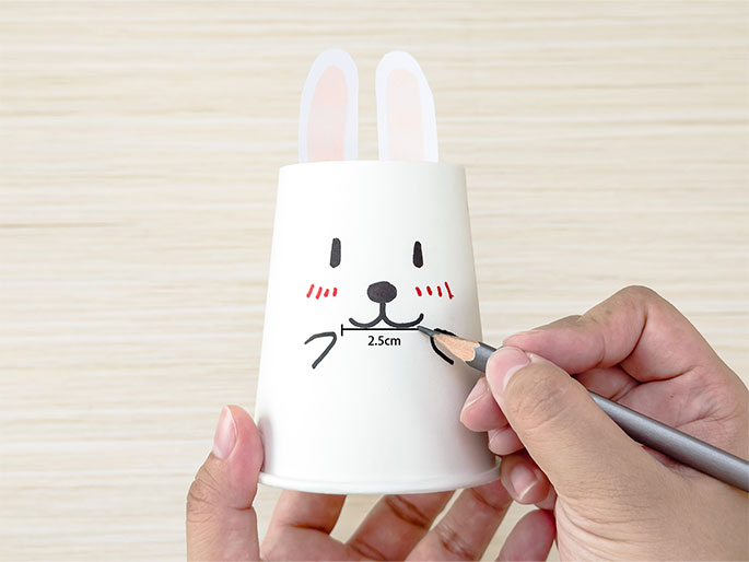 Two hands are working on a bunny-themed paper cup craft on a light wood background. The white cup has a drawn face with rosy cheeks, simple eyes, a nose, and a smiling mouth. Pink and white paper ears are attached to the rim. A hand holding a pencil is drawing a horizontal line 2.5cm below the mouth, likely to mark a cutting line or another element of the design. The image shows a step in creating a DIY bunny cup.
