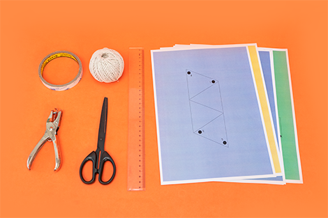 Arranged on a vibrant orange background are craft supplies: sticky tape, string, a hole punch, scissors, a ruler, and coloured sheets of paper printed with geometric shapes.