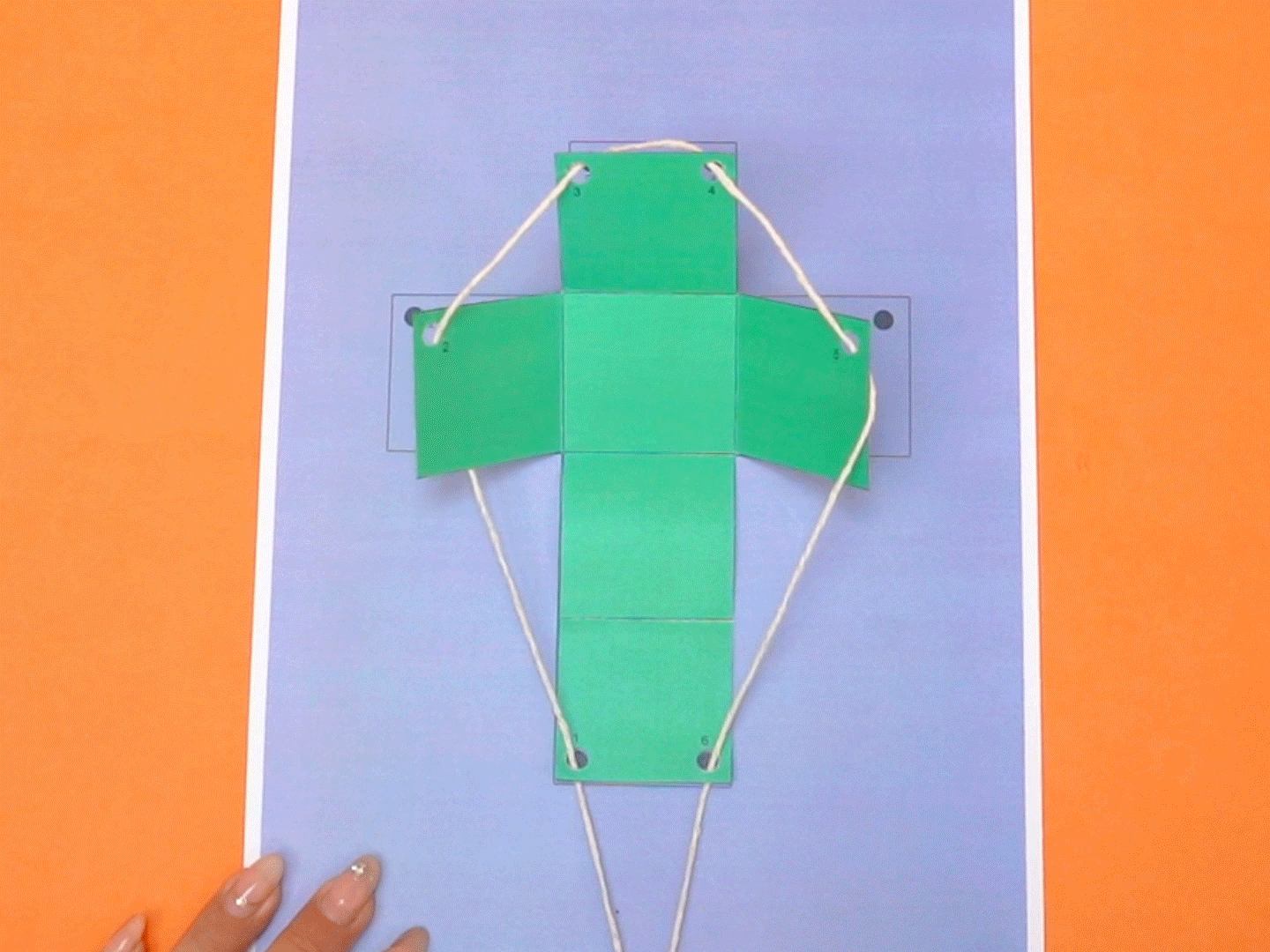 A partially constructed green paper cube, with string threaded through holes, is laid flat on a lavender-coloured piece of paper, which sits on an orange surface. A hand with long, decorated nails is visible at the bottom left.