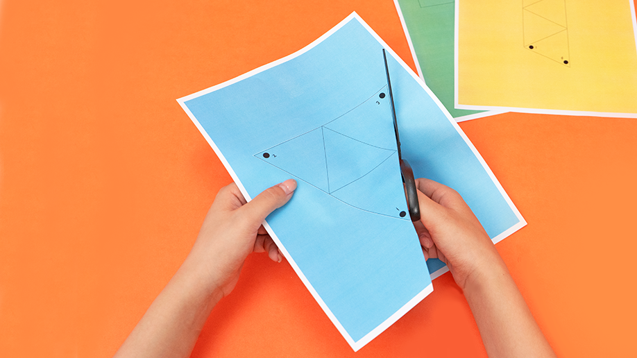 Hands use scissors to cut a light blue piece of paper, marked with a triangular outline, against a bright orange background; other coloured papers are visible in the top right corner.