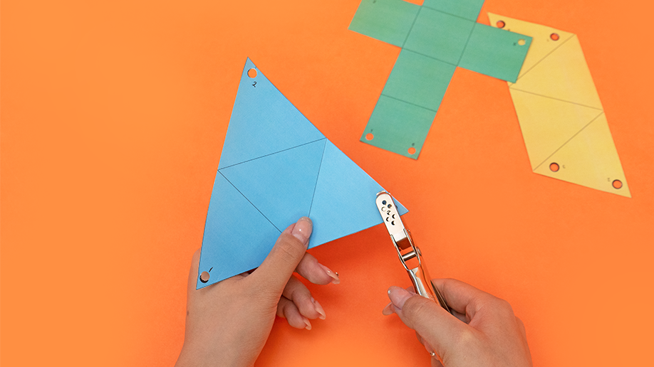 Two hands, one holding a light blue paper triangle and the other a hole punch, are shown against a bright orange background with other coloured shapes visible in the top right.