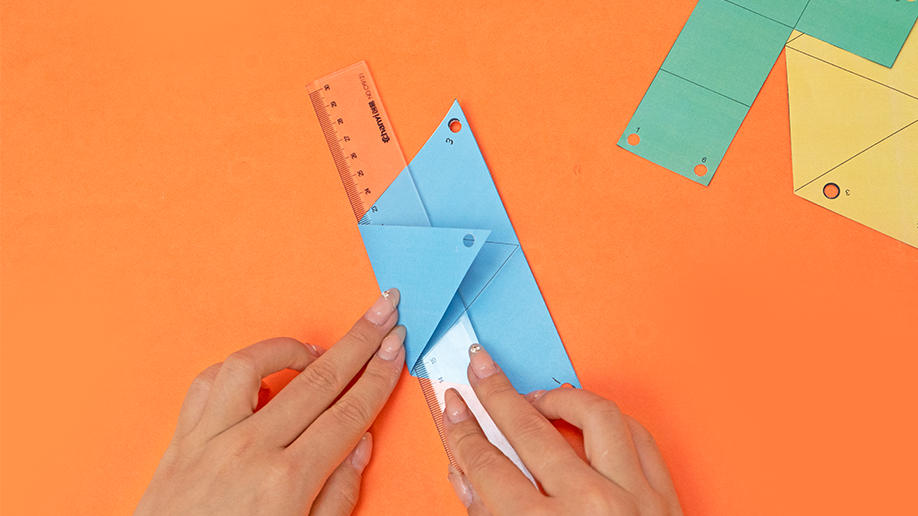 Two hands fold a light blue paper triangle along a crease, using a clear plastic ruler for accuracy, against a bright orange background; other coloured shapes are visible in the top right.