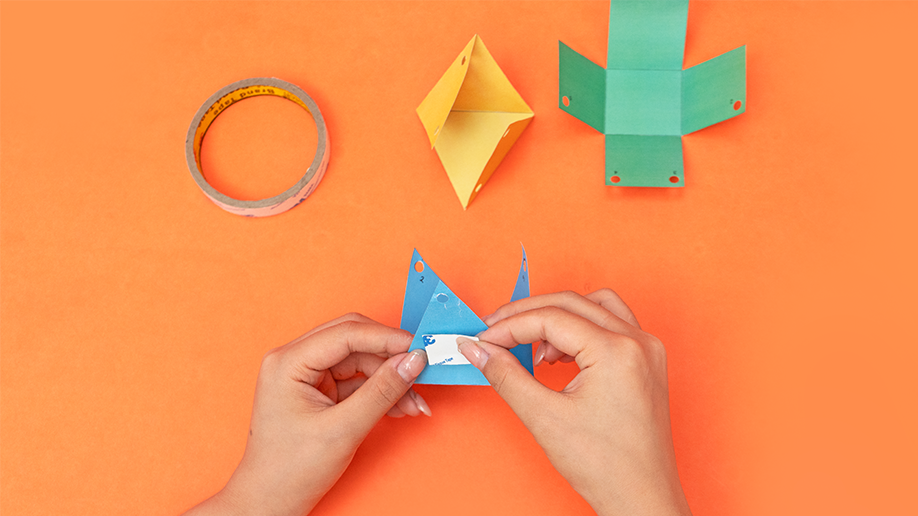 Two hands assemble a light blue paper shape, applying double-sided tape to one edge, against a vibrant orange background, with a roll of tape and other assembled coloured shapes visible.