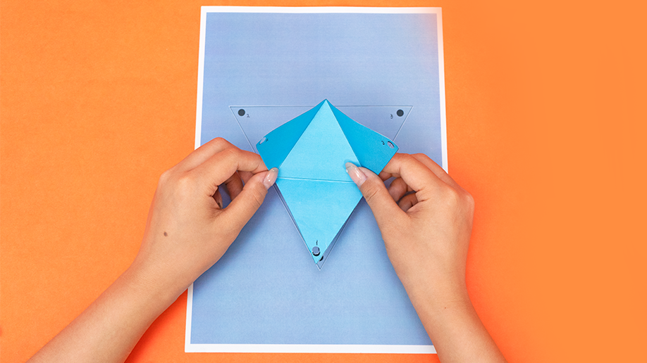 Two hands hold and shape a light blue paper tetrahedron over a lavender piece of paper on an orange surface.