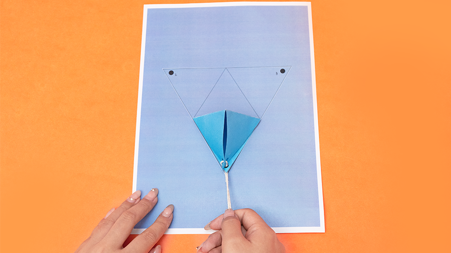 Two hands tie string around the bottom point of a light blue paper tetrahedron, which rests on a lavender piece of paper on an orange surface.