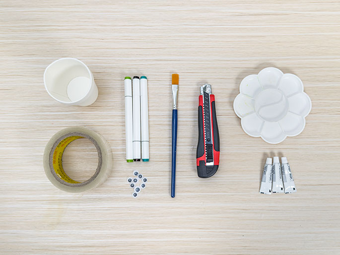 A paper cup, markers, a paintbrush, a utility knife, a paint palette, small tubes of paint, a roll of clear tape, and googly eyes are arranged on a light wood-grain surface.