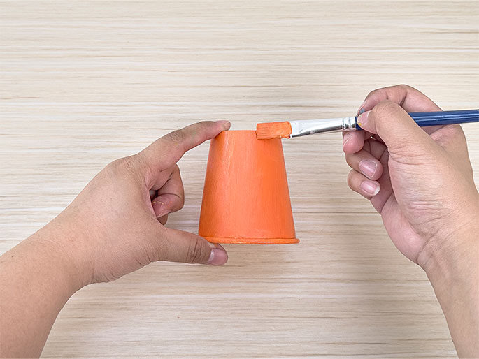 Two hands paint a small paper cup orange using a paintbrush on a light wood-grain surface.