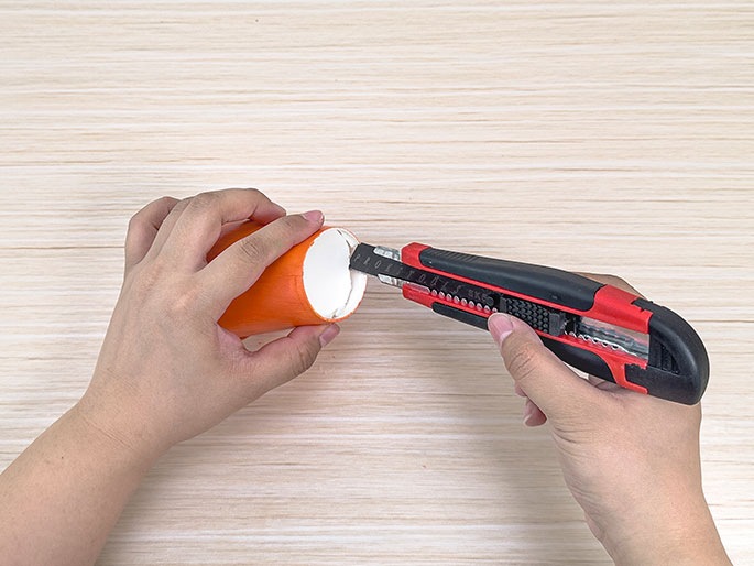 Two hands use a red and black utility knife to cut the rim of a small, orange paper cup on a light wood-grain surface.