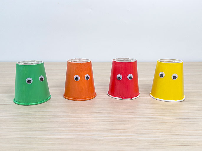 Four small paper cups, painted green, orange, red, and yellow, and decorated with googly eyes, are arranged in a row on a light wood-grain surface.