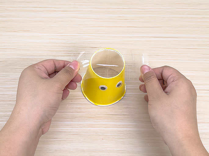 Two hands hold a piece of clear plastic wrap over a small, yellow paper cup with googly eyes on a light wood-grain surface.