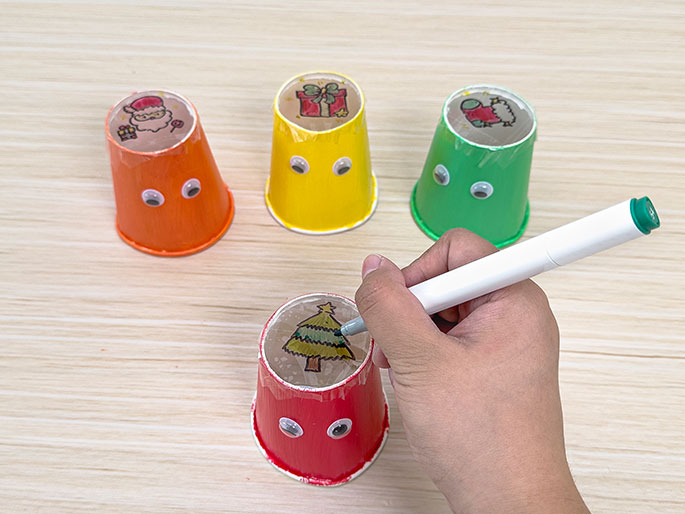 A hand uses a green marker to colour a drawing of a Christmas tree on plastic wrap covering a small, red paper cup with googly eyes, with three other similar cups, decorated with drawings of Santa Claus, a present, and a Christmas stocking, in the background on a light wood-grain surface.