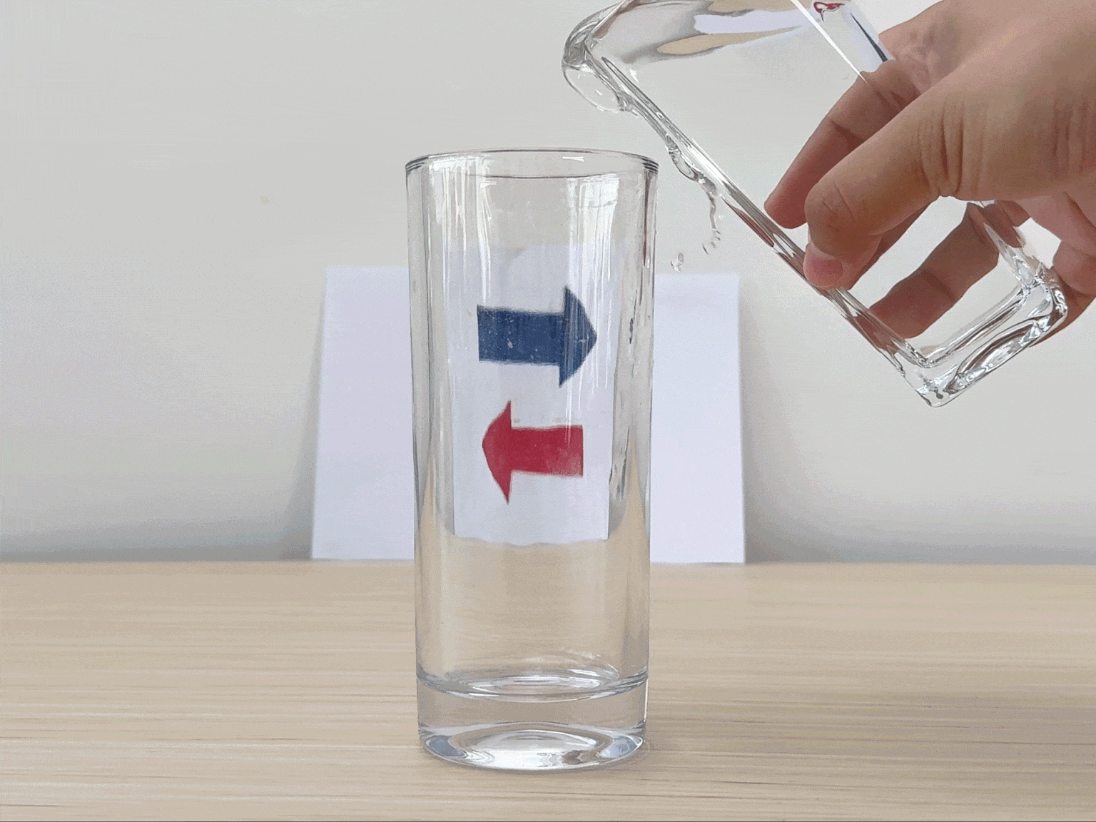 A clear glass sits on a light wooden table with a piece of white paper positioned behind it. The paper features two arrows: a blue arrow pointing to the right and a red arrow pointing to the left. A hand pours water from a clear glass pitcher into the empty glass. As the glass fills, the arrows appear to change direction due to the refraction of light through the water. This video demonstrates a simple science experiment illustrating the principles of light refraction.