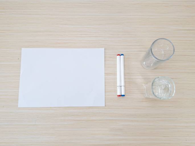 Arranged on a light wood-grain table are several items, suggesting preparation for an activity or demonstration. A blank white sheet of paper lies on the left. Two white markers, one with a red cap and the other with a blue cap, are positioned next to the paper. Two clear drinking glasses are on the right; one is empty and the other contains a small amount of water.