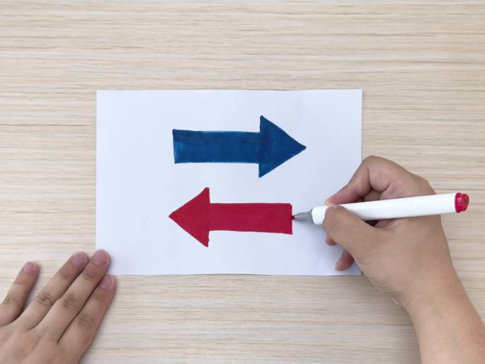 On a light wood-grain table, a piece of white paper displays a blue arrow pointing to the right. A hand holding a red marker is drawing a second arrow directly beneath the blue one, pointing to the left. Another hand is positioned near the bottom left corner of the paper. This scene likely depicts someone creating directional arrows, perhaps for a presentation or a game.
