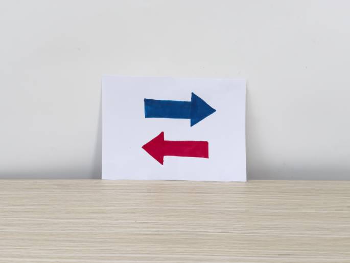 Against a plain white background, a piece of white paper rests on a light wood-grain surface. Drawn on the paper are two arrows: a blue arrow pointing to the right and a red arrow pointing to the left, positioned one above the other. This simple image conveys a sense of direction or opposing choices.