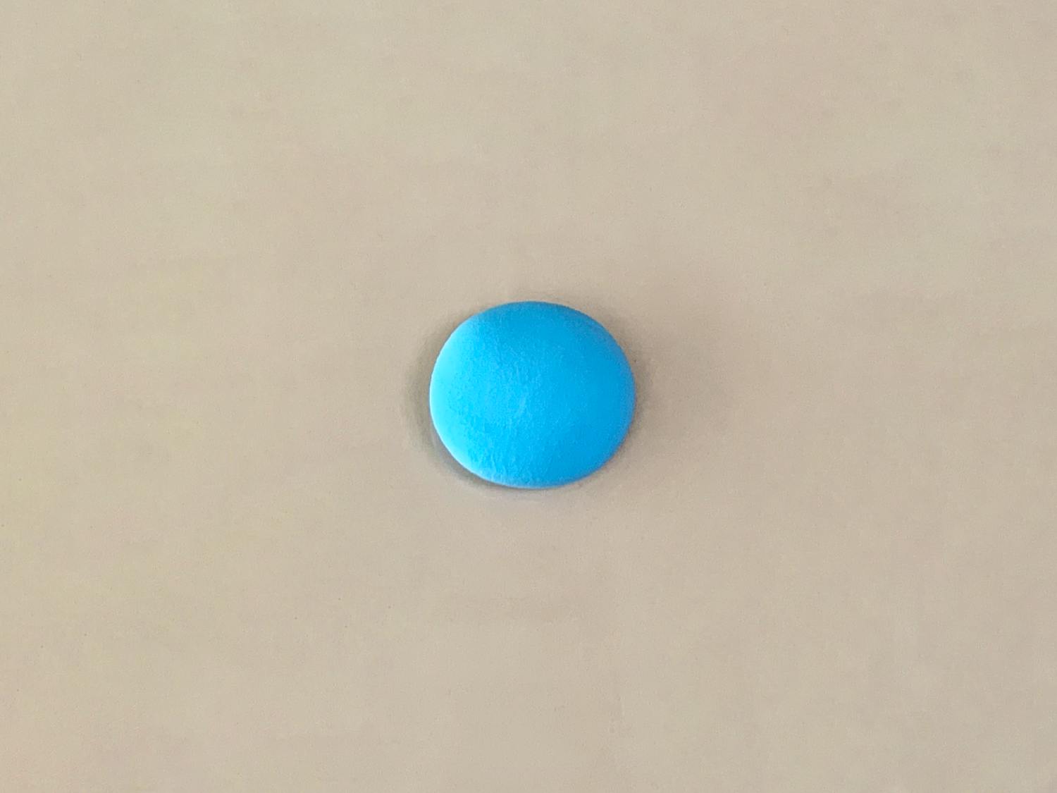 A small, flattened oval of bright blue playdough rests on a plain, light beige surface.