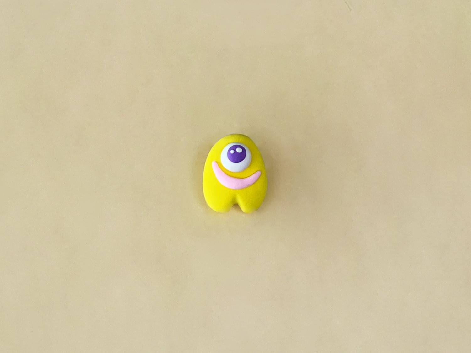 A small, bright yellow playdough figure, shaped like a rounded body with two small legs, has a purple googly eye and a pink smile, and sits on a light beige surface.