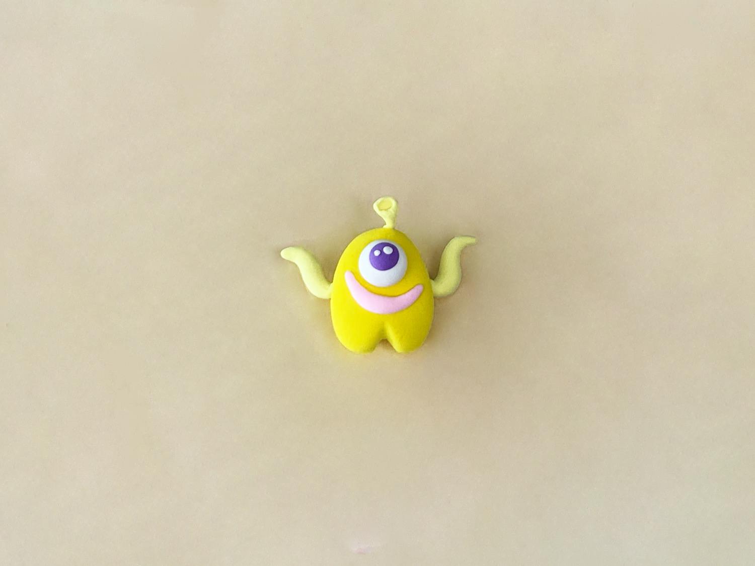 A small, bright yellow playdough figure, shaped like a rounded body with two small legs, has a purple googly eye, a pink smile, two small, light yellow arms, and a small, light yellow antenna, and sits on a light beige surface.