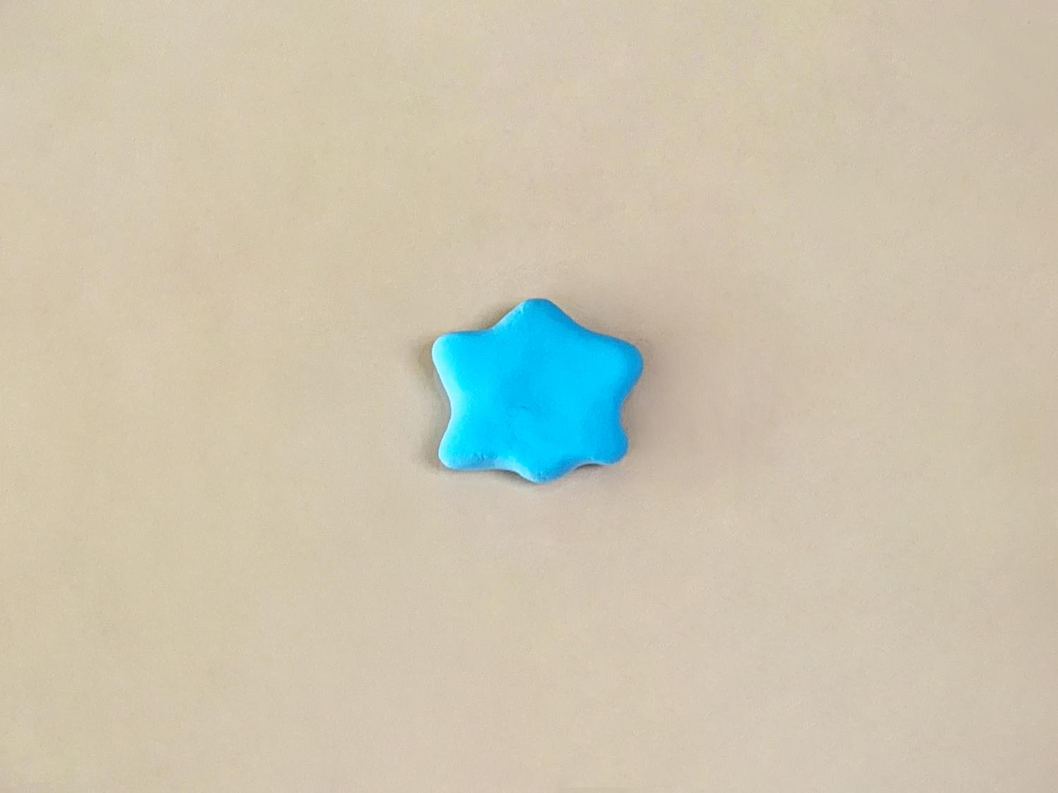 A small, five-pointed star made of bright blue playdough sits on a plain, light beige surface.