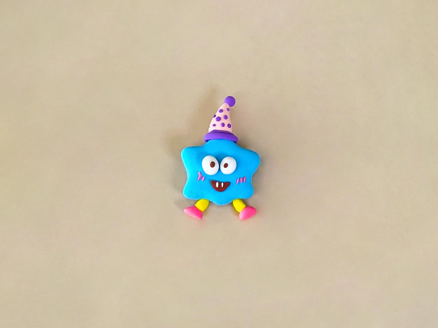 A small, light blue, star-shaped playdough figure with googly eyes, a smiling mouth with fangs, yellow legs, pink feet, and a light purple and pink polka-dotted party hat sits on a light beige surface.