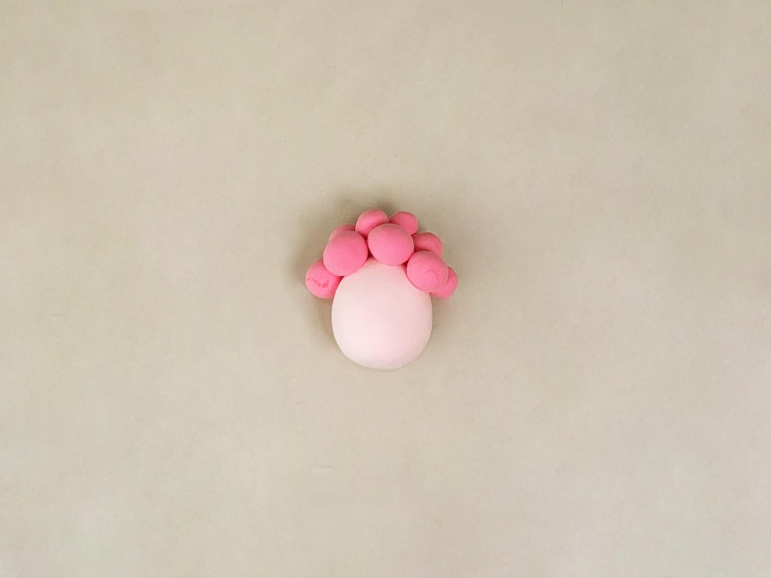 A pale pink ball of playdough, topped with a semi-circle of smaller, bright pink balls, sits on a plain, light beige surface.