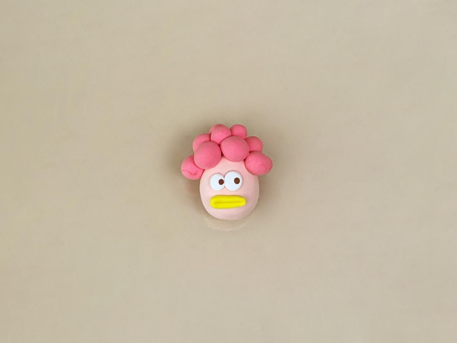 A small figure made of pale pink playdough, with bright pink hair, googly eyes, and a yellow beak, sits on a plain, light beige surface.