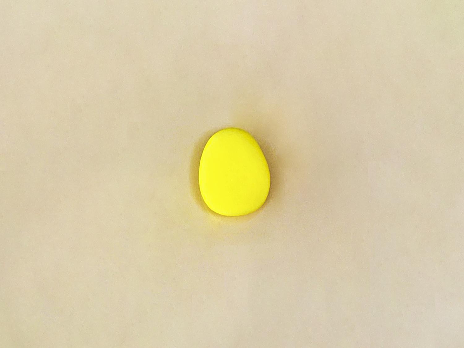 A small, flattened oval of bright yellow playdough rests on a plain, light beige surface.