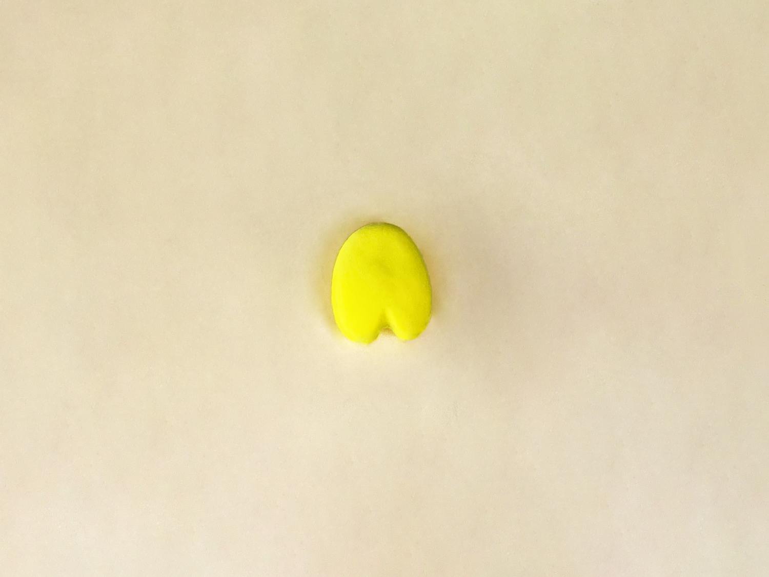 A small, rounded, bright yellow piece of playdough, pinched at the bottom to resemble a pair of legs or the bottom of a body, sits on a plain, light beige surface.