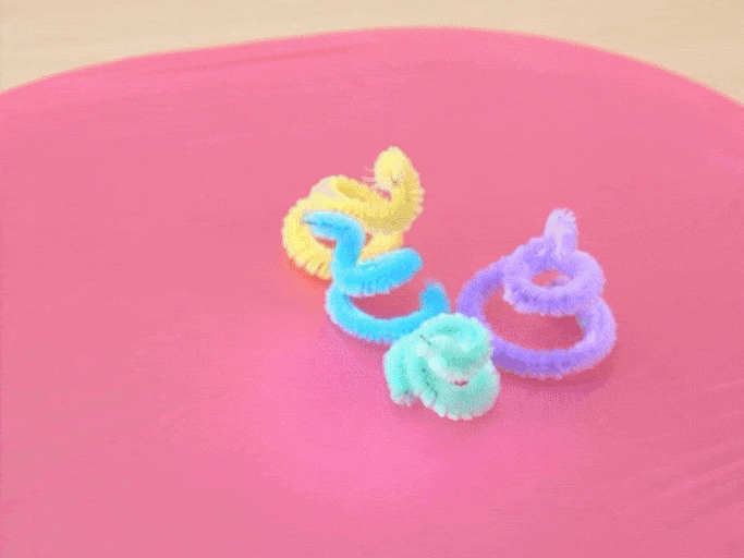 Several colorful pipe cleaners, shaped into spirals and rings, rest on a bright pink surface. The pipe cleaners are yellow, purple, light green, and light blue. They shift slightly, suggesting they might be on a vibrating surface or being gently moved. The video showcases a playful and simple craft project, likely aimed at children.