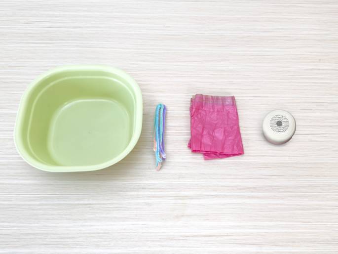 Laid out on a light wood-grained surface are a few items, likely for a craft or science activity. A pale green, shallow rectangular basin sits on the left. In the center is a small bundle of pastel-colored pipe cleaners. To the right, a crumpled piece of bright pink tissue paper lies next to a small, round, silver object with a perforated top, possibly a speaker or diffuser.