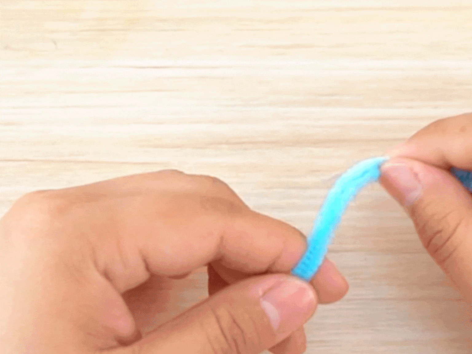 Two hands manipulate a light blue pipe cleaner against a blurred light wooden background. The fingers gently bend and shape the fuzzy wire, likely demonstrating a crafting technique or the initial steps in making a pipe cleaner creation.