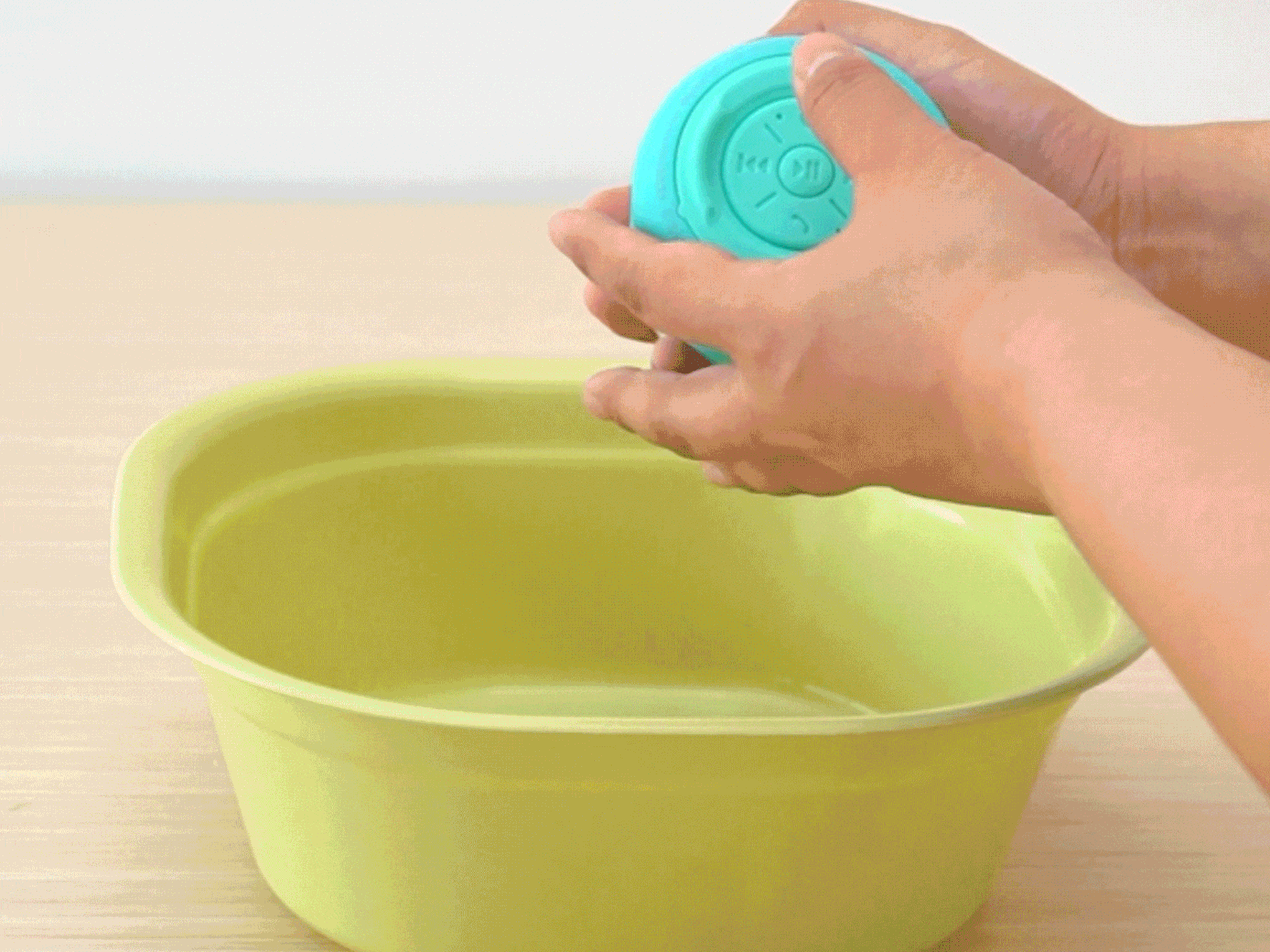 Hands hold a small, round, teal Bluetooth speaker over a pale green plastic basin on a light wood-grain table. The speaker has visible control buttons. The hands appear to be about to place the speaker into the basin, possibly demonstrating its waterproof capabilities or preparing it for an unusual use.