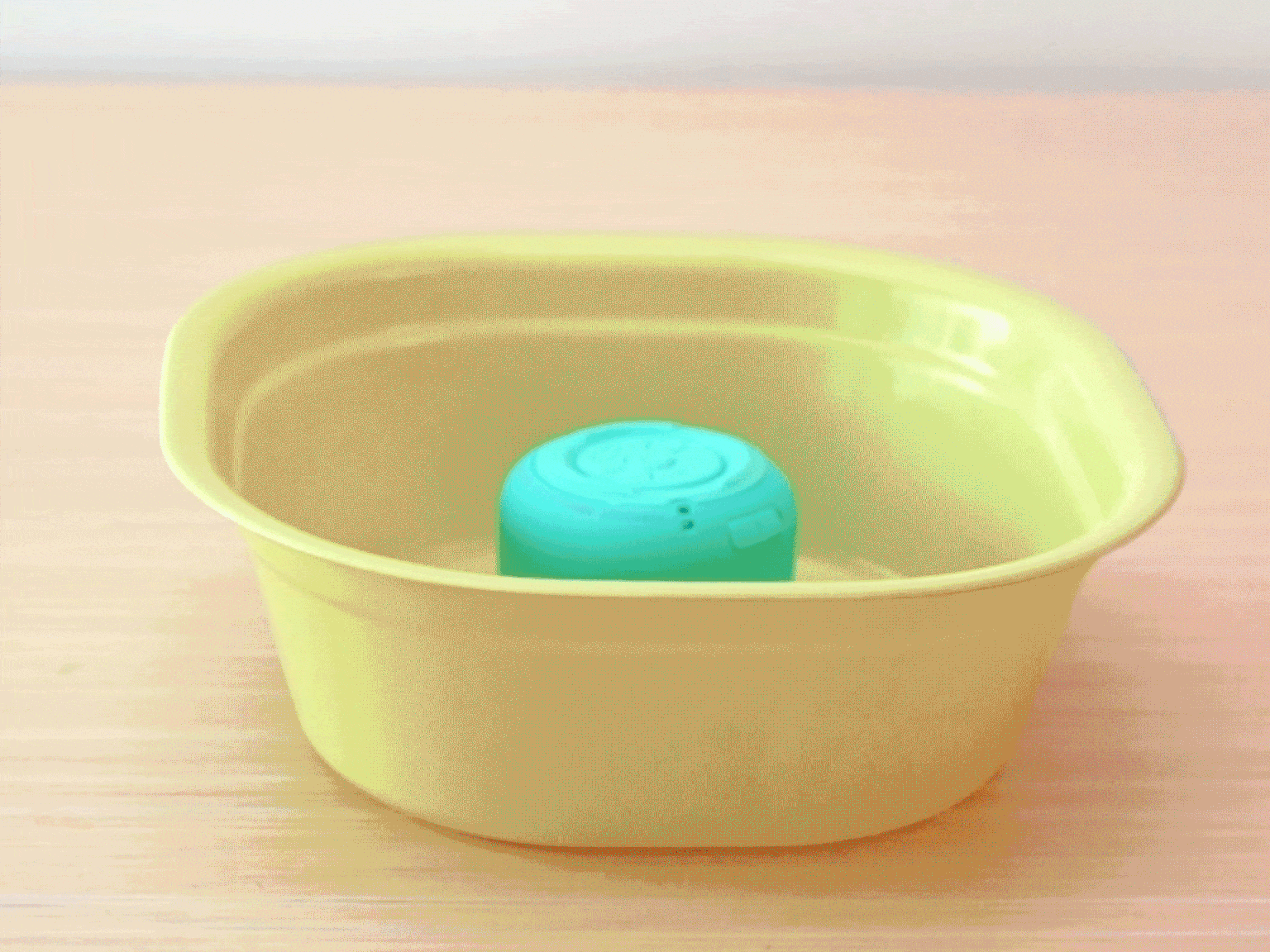 A small, round, teal Bluetooth speaker sits in the bottom of a pale green plastic basin on a light wood-grain table. The speaker is dry and appears to be ready for use, possibly demonstrating its waterproof nature or being used in an unconventional way.