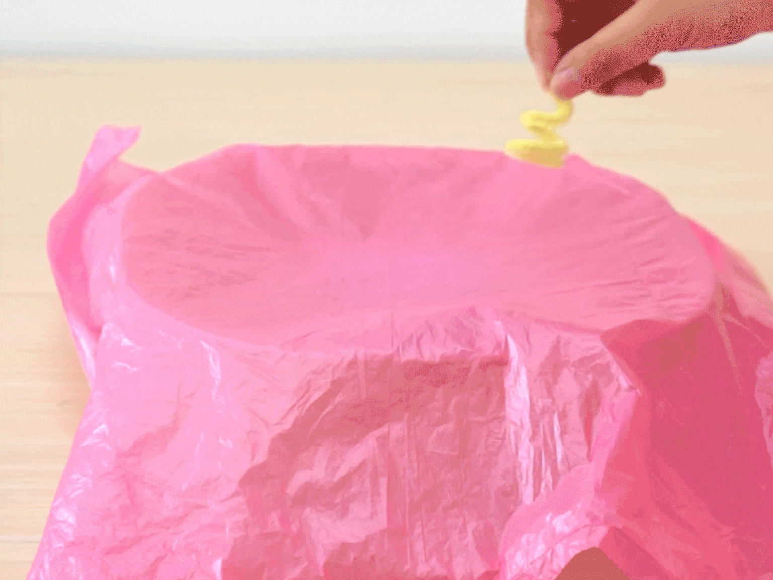 A hand holds a twisted yellow pipe cleaner above a piece of crumpled bright pink tissue paper on a light wooden surface. The hand appears to be about to place or drop the pipe cleaner onto the tissue paper, possibly as part of a craft project or demonstration.
