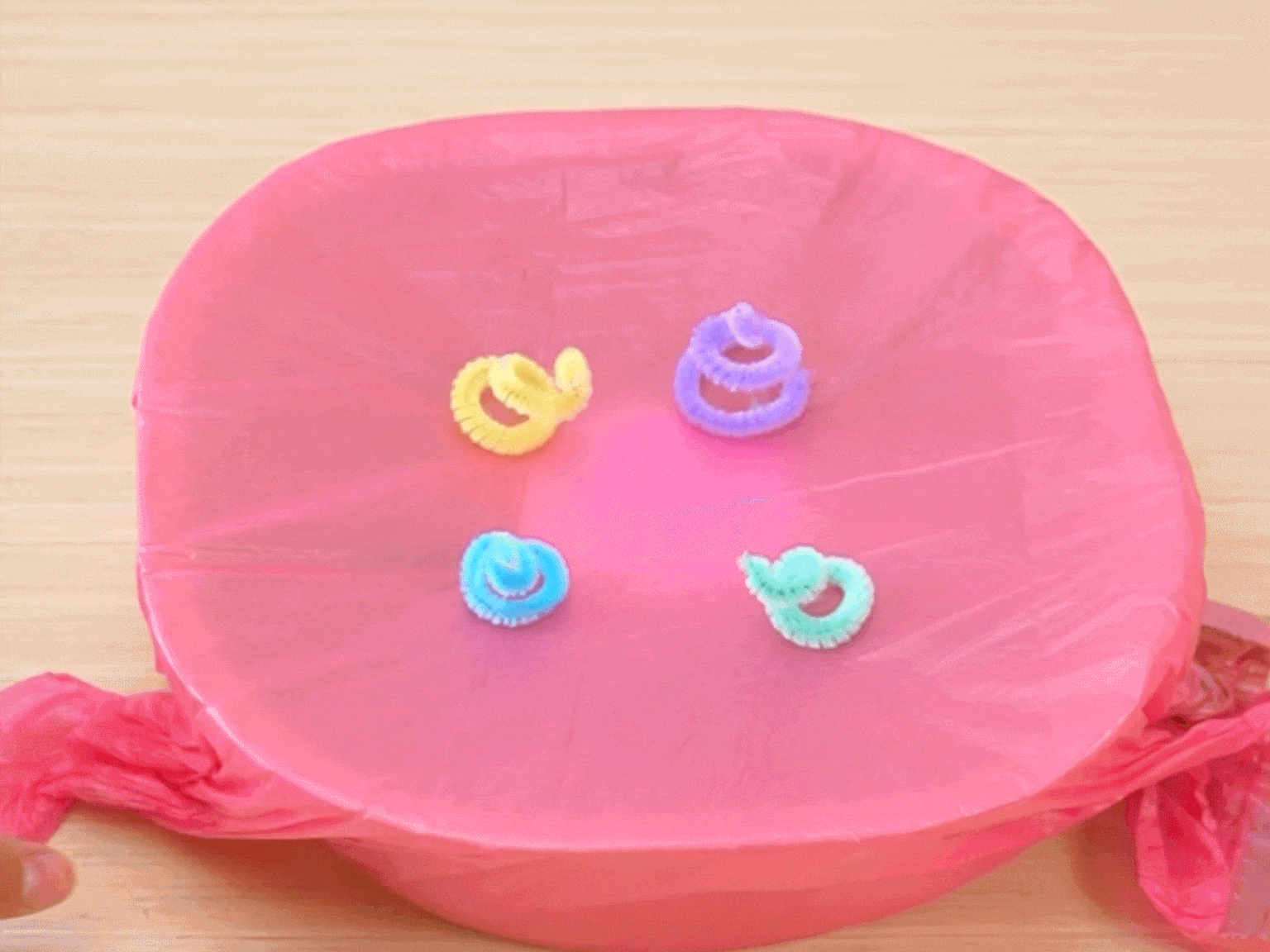 Several small, twisted pipe cleaners in yellow, purple, light blue, and light green are placed on a stretched piece of bright pink tissue paper. The tissue paper is positioned over a container or opening, and a hand is visible at the bottom left corner, likely manipulating the tissue paper or the setup. This suggests a craft activity or experiment in progress, possibly involving static electricity or the pipe cleaners' interaction with the tissue paper.