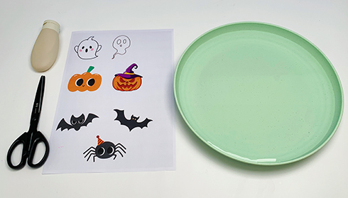 Gather your spooky supplies! A sheet of cute Halloween character templates, a pair of scissors, a bottle of glue, and a pale green plate are all laid out, ready for some frightfully fun crafting. Let's get those spooky creations underway!