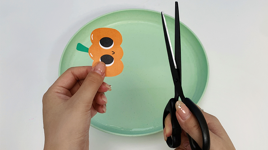 Getting those spooky shapes ready! A pair of hands carefully holds a paper pumpkin, while another hand holds scissors, poised to make a precise cut. It's all about those spooky details!