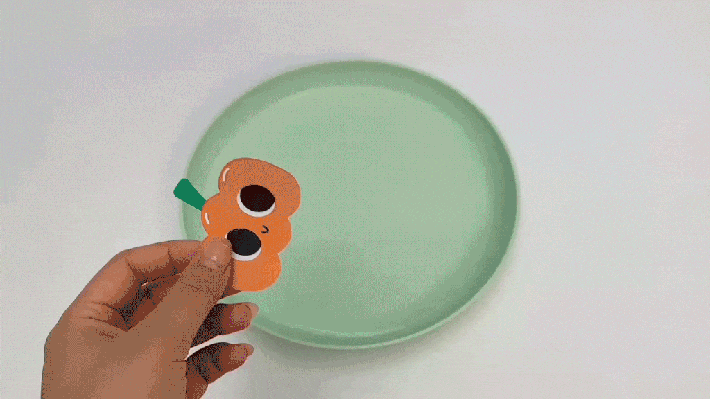 Adding a touch of spooky charm! A hand carefully places a cute paper pumpkin onto a pale green plate, its friendly face adding a touch of Halloween whimsy. It's looking rather splendid!