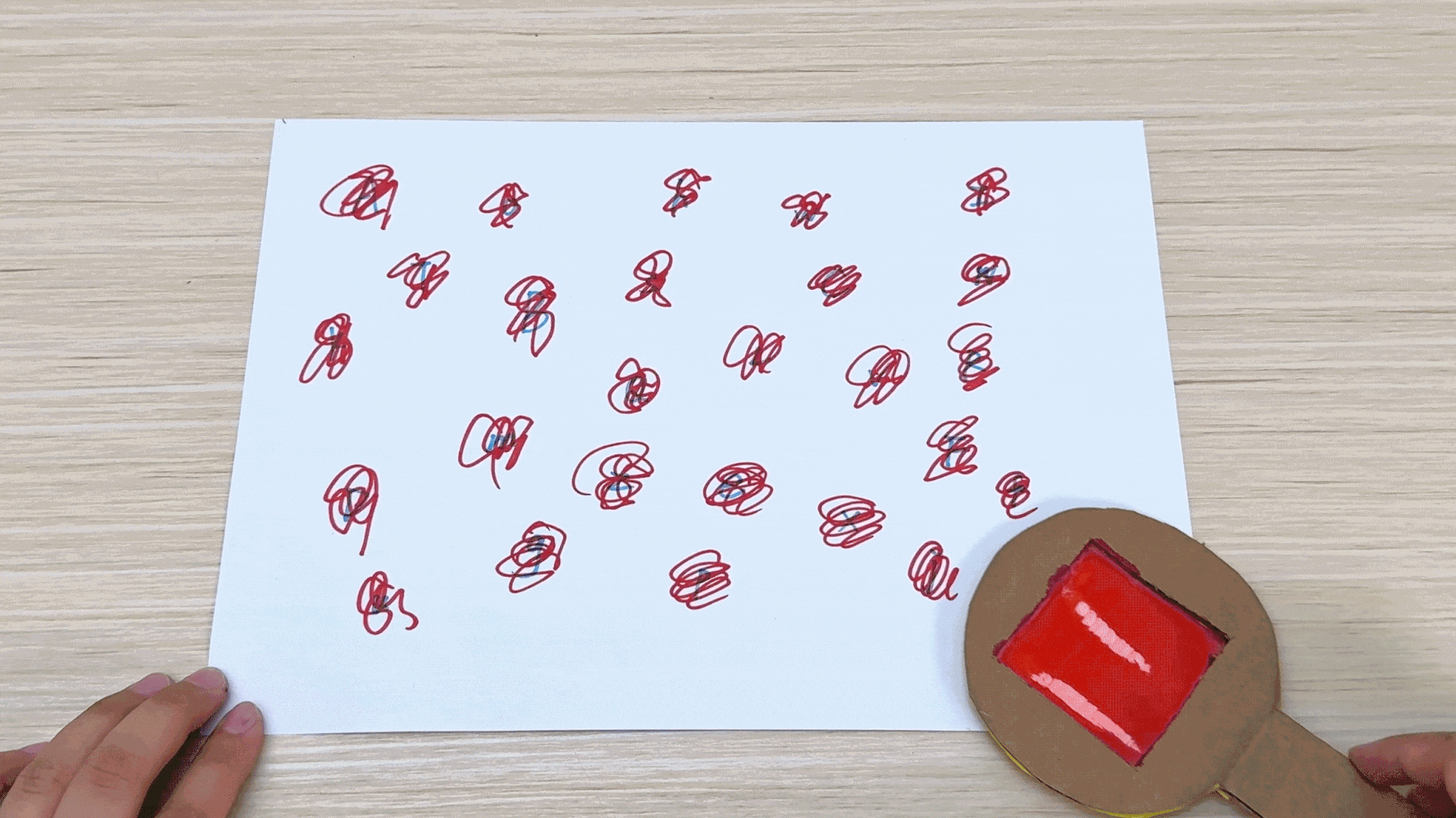 Hands manipulate a homemade magnifying glass made from cardboard and red cellophane. The magnifying glass is moved across a piece of paper with numerous red scribbles, demonstrating a simple optical device.