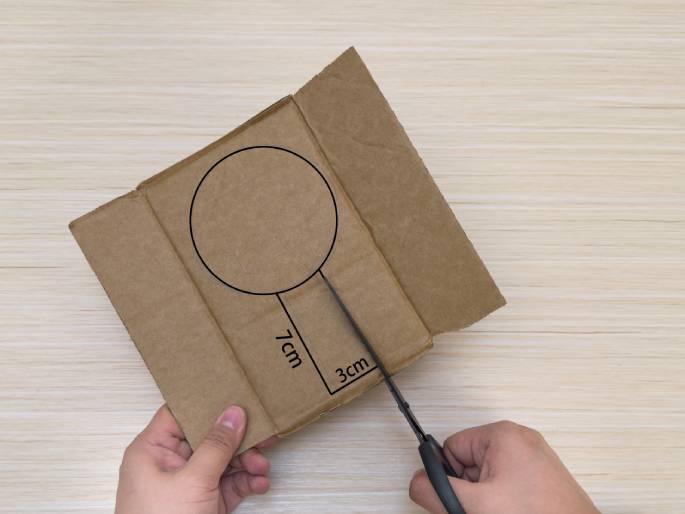 Two hands are cutting a piece of cardboard on a light wood-grain table. A circle and a connected rectangle have been drawn on the cardboard, resembling a lollipop or paddle shape. The rectangle has 7cm written along its length and 3cm written along its shorter base. Black scissors held in the right hand are cutting along the bottom edge of the rectangular shape. This image appears to be a step in a craft or DIY project.