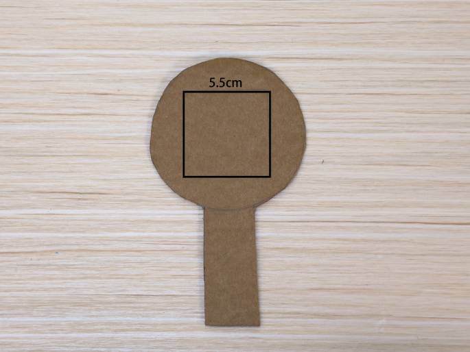 A lollipop-shaped piece of cardboard rests on a light wood-grain table. A square is drawn within the circular top part of the shape, with the notation 5.5cm above it, indicating the square's side length. This cardboard cutout likely serves as a template or component in a craft or DIY project.