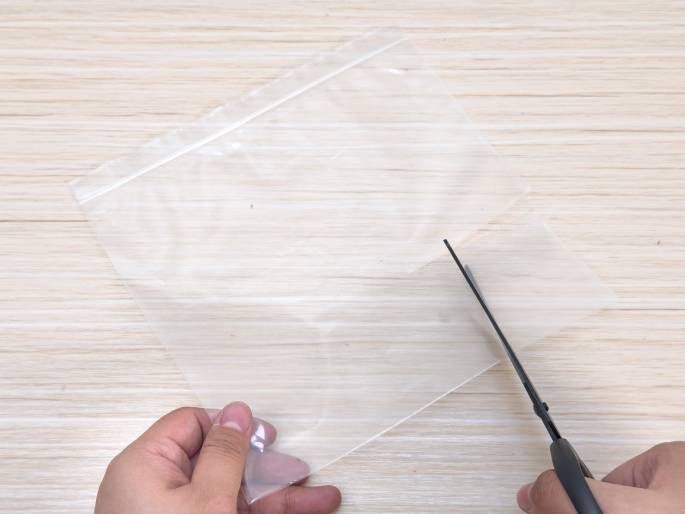Two hands are shown cutting a clear plastic bag or sheet on a light wood-grain table. The left hand holds the plastic flat while the right hand uses black scissors to cut along one edge. The plastic appears to be a resealable bag, as a zip-top closure is visible along the top edge. This image likely depicts a step in a craft or DIY project.