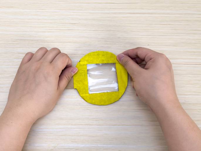 Two hands hold a circular piece of cardboard with a rectangular window cut into it on a light wood-grain table. The cardboard is covered with yellow tape, and a piece of clear plastic is affixed over the window, creating a transparent viewing area. The hands are holding the edges of the cardboard, likely demonstrating a stage in a craft or DIY project.