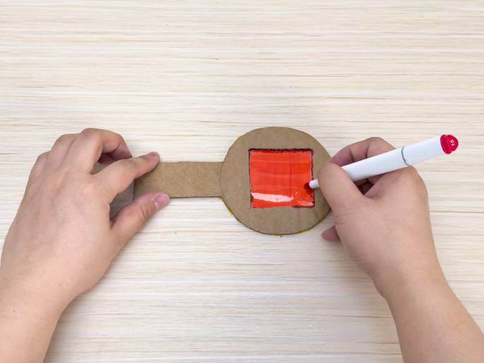 Two hands are working with a lollipop-shaped piece of cardboard on a light wood-grain table. The circular part of the cardboard has a square window covered with a piece of clear plastic. The right hand holds a white marker with a pink cap and is coloring the plastic red. The left hand holds the cardboard steady. This image likely shows a step in creating a homemade color filter or a similar craft project.