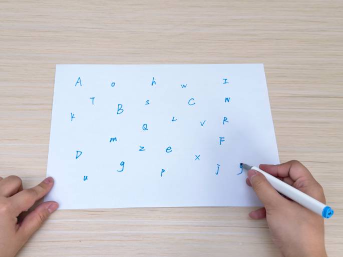 On a light wood-grain table, two hands interact with a white sheet of paper. The paper has various uppercase and lowercase letters written on it in blue ink, scattered seemingly randomly across the surface. The right hand holds a white marker with a light blue cap and is in the process of writing on the paper. The left hand rests on the bottom left corner of the paper. The scene suggests an activity involving writing, perhaps a word game or a learning exercise.