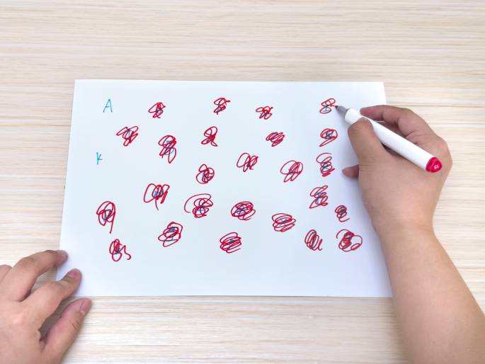 Two hands and a piece of paper are shown on a light wood-grain table. The paper is filled with numerous red scribbles and swirls, each with a small blue mark near its center. The right hand holds a white marker with a pink cap and appears to be adding more red scribbles to the paper. The left hand rests on the bottom left corner of the paper. The image likely depicts a game, a creative activity, or a learning exercise involving visual tracking or pattern recognition. A blue A and a blue k are written in the top left corner.