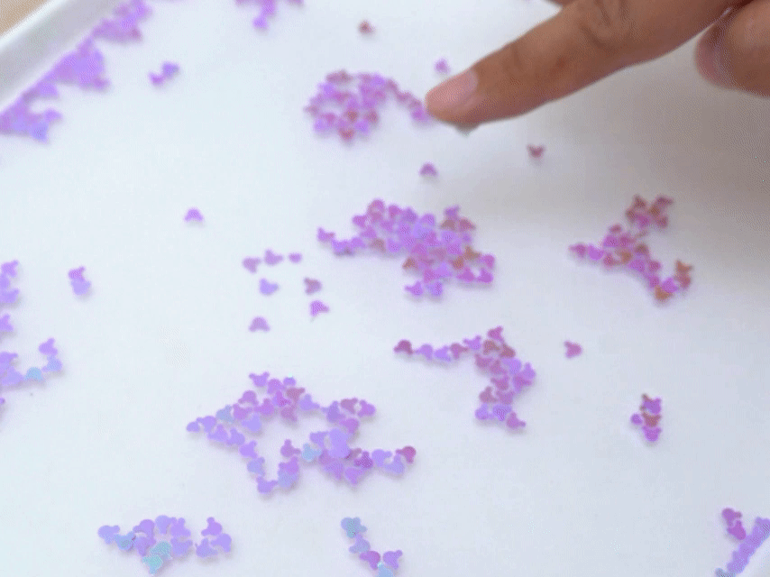 A finger traces a path through iridescent purple Mickey Mouse-shaped confetti on a white surface. The confetti is spread out, and the finger moves some of it aside, creating a clear space.